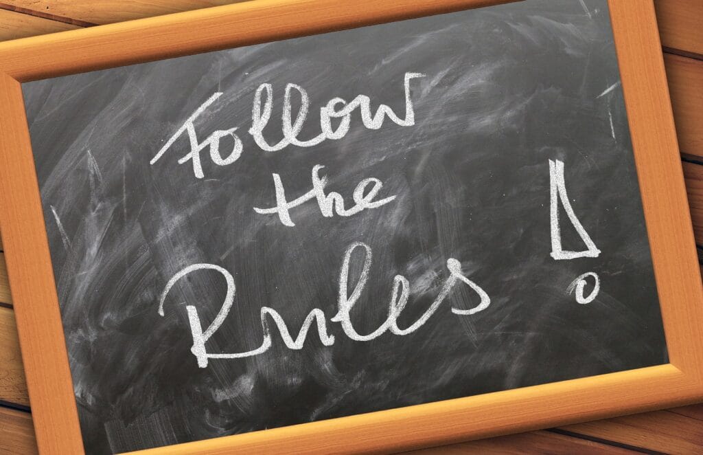 The image shows a chalkboard with the words "Follow the Rules!" written in white chalk. The handwriting is casual and the message is emphasized by an exclamation mark at the end. The chalkboard is framed with a wooden border and appears to be placed on a wooden surface, suggesting a friendly reminder or instruction to follow guidelines.