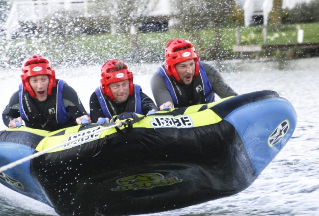 The image shows three men riding a large inflatable water tube while being towed at high speed across the water. They are wearing red helmets and life vests for safety, gripping tightly onto the handles of the tube. The expressions on their faces show a mix of excitement and concentration, with water splashing around them as the tube skims across the surface. The tube is branded with the name “Jobe,” and the scene captures the thrilling, fast-paced nature of watersports.