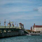 The Bridge of Lions in St. Augustine, Florida, stretching across the Matanzas River with its distinctive green arches and red-roofed towers. The historic downtown skyline, including iconic Spanish-style buildings, is visible in the background on a clear day.