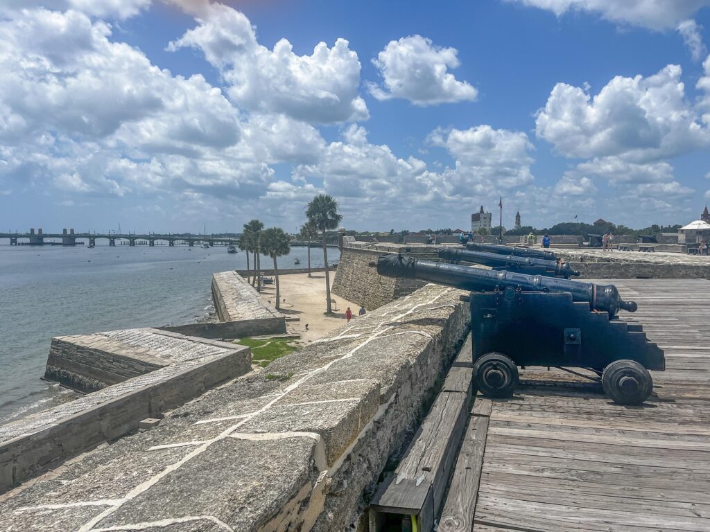 A historic view from the Castillo de San Marcos in St. Augustine, Florida, featuring old black cannons lined up along the stone fortress walls overlooking the Matanzas River. The Bridge of Lions and the downtown skyline, including Spanish-style buildings, are visible in the background under a bright blue sky with scattered clouds.